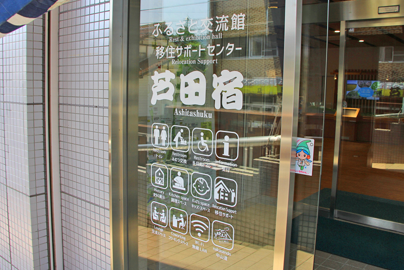 ガラスの入り口ドアに「ふるさと交流館 移住サポートセンター 芦田宿」の文字と、トイレ、おむつ交換代など12種類のピクトグラムが表示されている正面ドアの写真