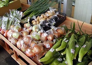 とうもろこし、玉ねぎ、ネギなどの野菜が棚に並べられている農産物直売所の写真