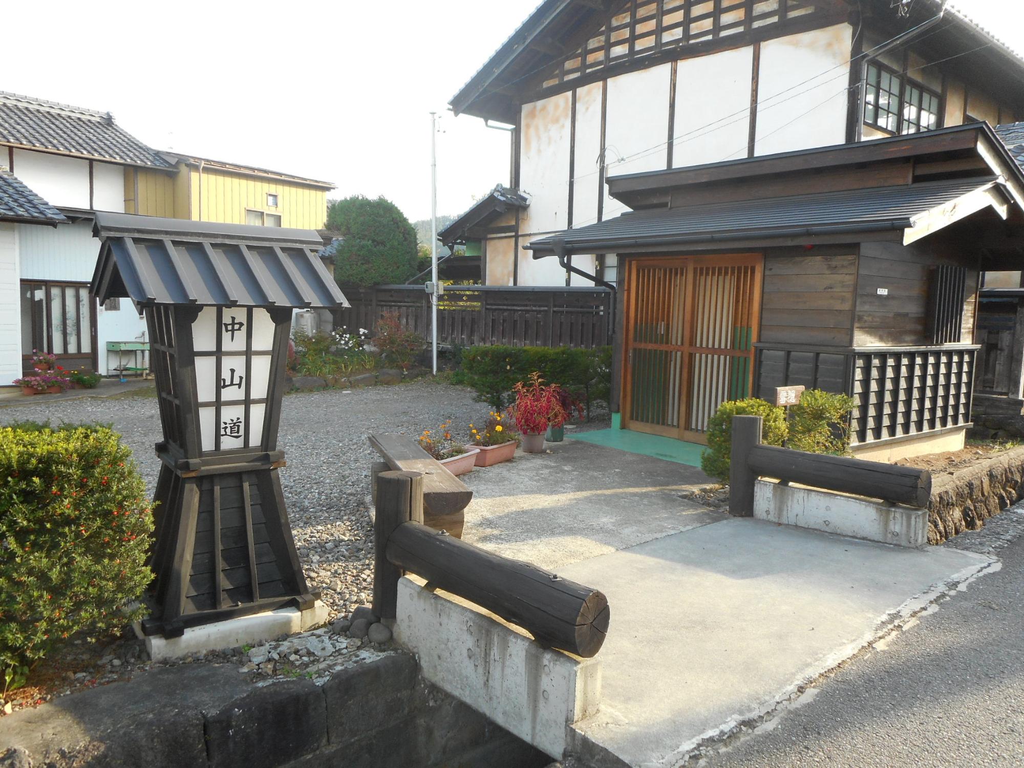 左側に「中山道」と書かれた屋根付きの立看板があり、右側には格子戸の入口がある白い外壁の中山道 茂田井間の宿の外観写真