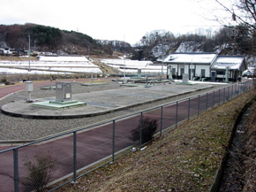 奥に平屋の建物があり、手前の広いスペースに長円形の形をした場所が見える茂田井浄化センターの外観写真