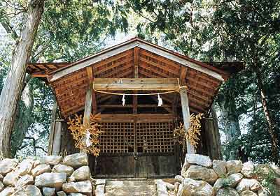 周りに木々が立っており、石が沢山積み重ねられている石垣の上にある古い木造の諏訪神社の外観写真