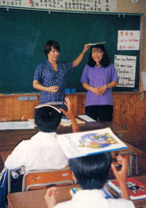 ALTの女性の先生が黒板の前に立ち、隣に立つ女性の頭の上に教科書を乗せて、子どもたちに英語指導を行う様子の写真
